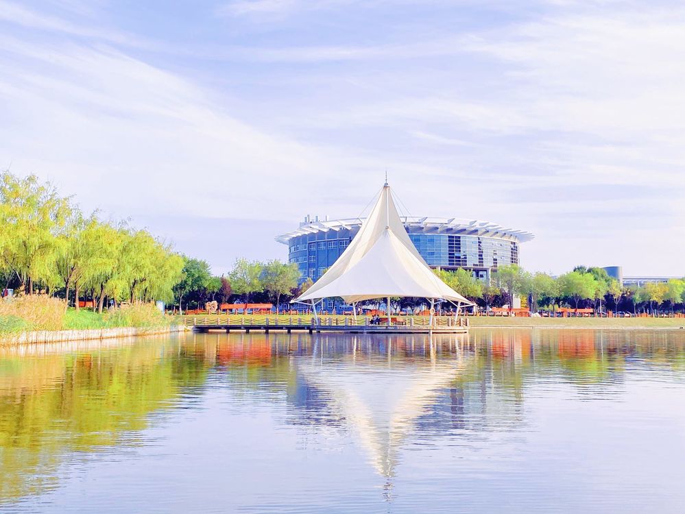 Lakeside pavilion with a white conical tent roof reflected in calm water, trees and modern building behind, blue and green tones